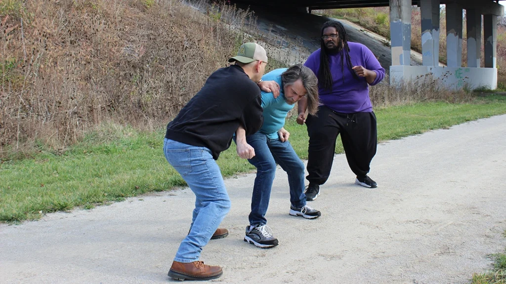 The author uses a controlling/dragging armbar and shoulder lock to keep his attacker off balance and in front of him—in the way of the larger attacker. Essentially, taking a hostage.